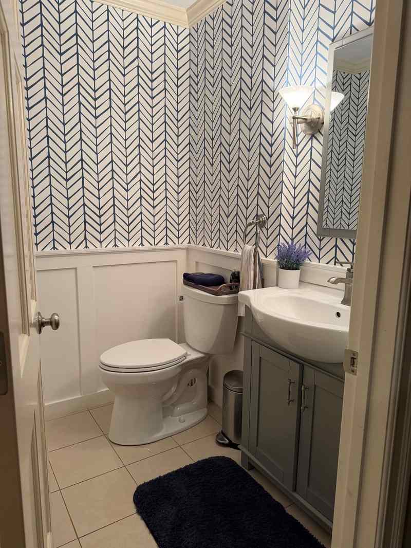 A finished powder room with herringbone wallpaper over wainscoting and a gray vanity