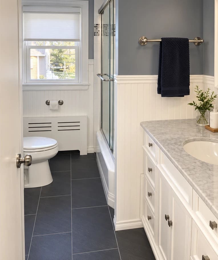 The same bathroom after the update, with white beadboard wainscoting, gray-blue walls, large dark floor tile, a new white vanity and a marble-look top