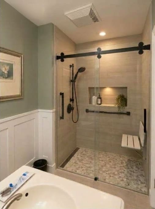 The same bathroom with a tiled walk-in shower, glass slider, wall niche, fold-down seat and white wainscoting